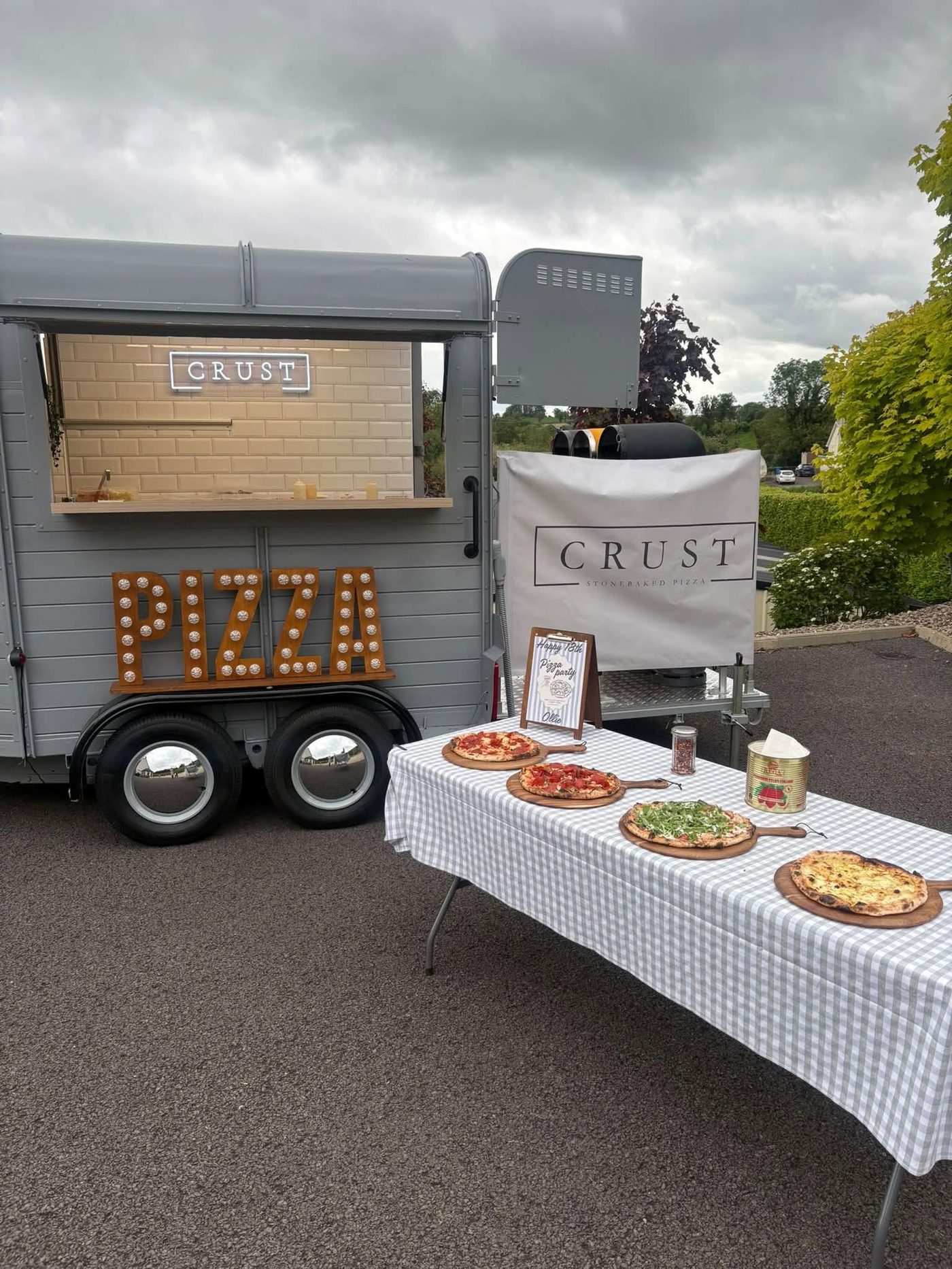 The Crust trailer set up at an event with fresh pizzas on serving boards